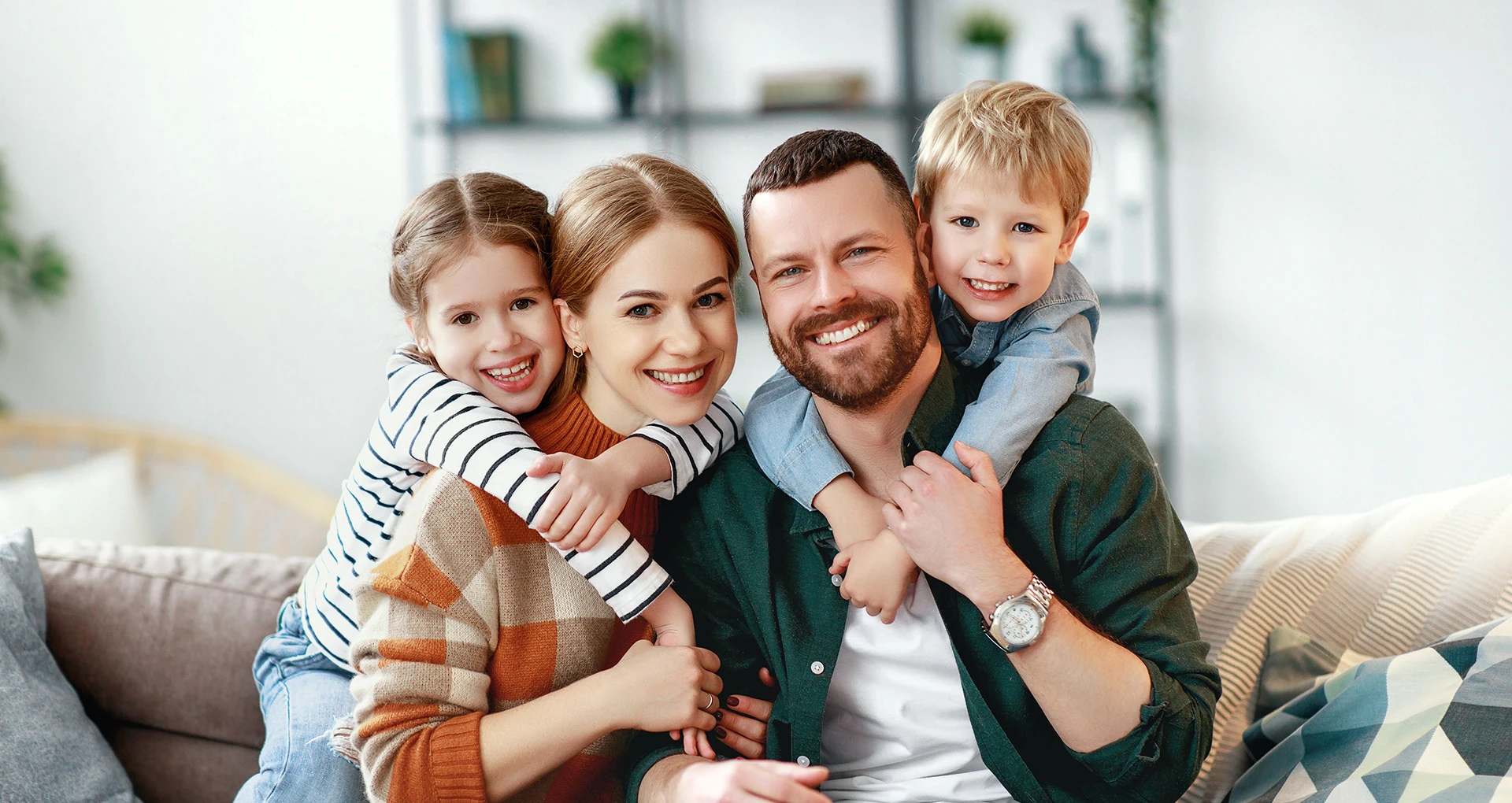 Eine Familie bestehend aus einem Mann, einer Frau und zwei Kindern hält sich in den Armen und strahlt lächelnd in Ihre Richtung.